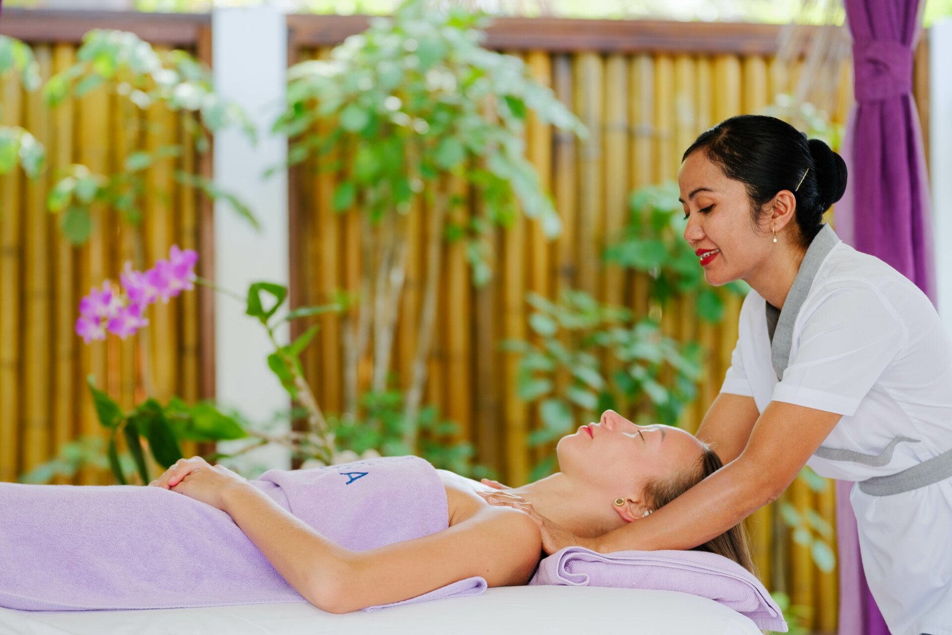 Behandlung im ELENA THE SPA Eine Entspannungsszene in einem Spa: Eine Frau in professioneller weißer Kleidung massiert den Nacken einer entspannt liegenden Kundin, die auf einer Liege mit lila Handtüchern bedeckt ist. Im Hintergrund ist eine Bambuswand sowie üppige grüne Pflanzen und einige lila Blumen zu sehen.