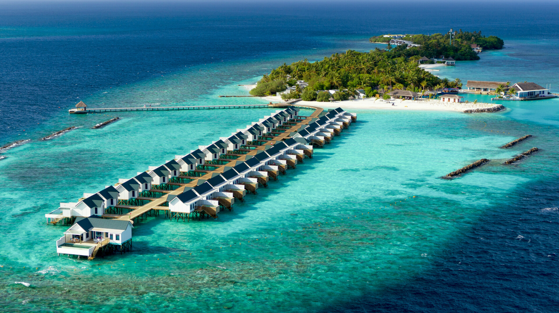 Insel von oben Luftaufnahme eines luxuriösen Inselresorts mit einer Reihe moderner Überwasser-Bungalows in lebhaften Farben, die sich auf kristallklarem, türkisfarbenem Wasser erstrecken. Eine Insel mit üppigem Grün und weißen Sandstränden umgibt die Bungalows, verbunden durch lange Stege. Das Bild zeigt ein tropisches Paradies, ideal für Erholung und Flucht aus dem Alltag.