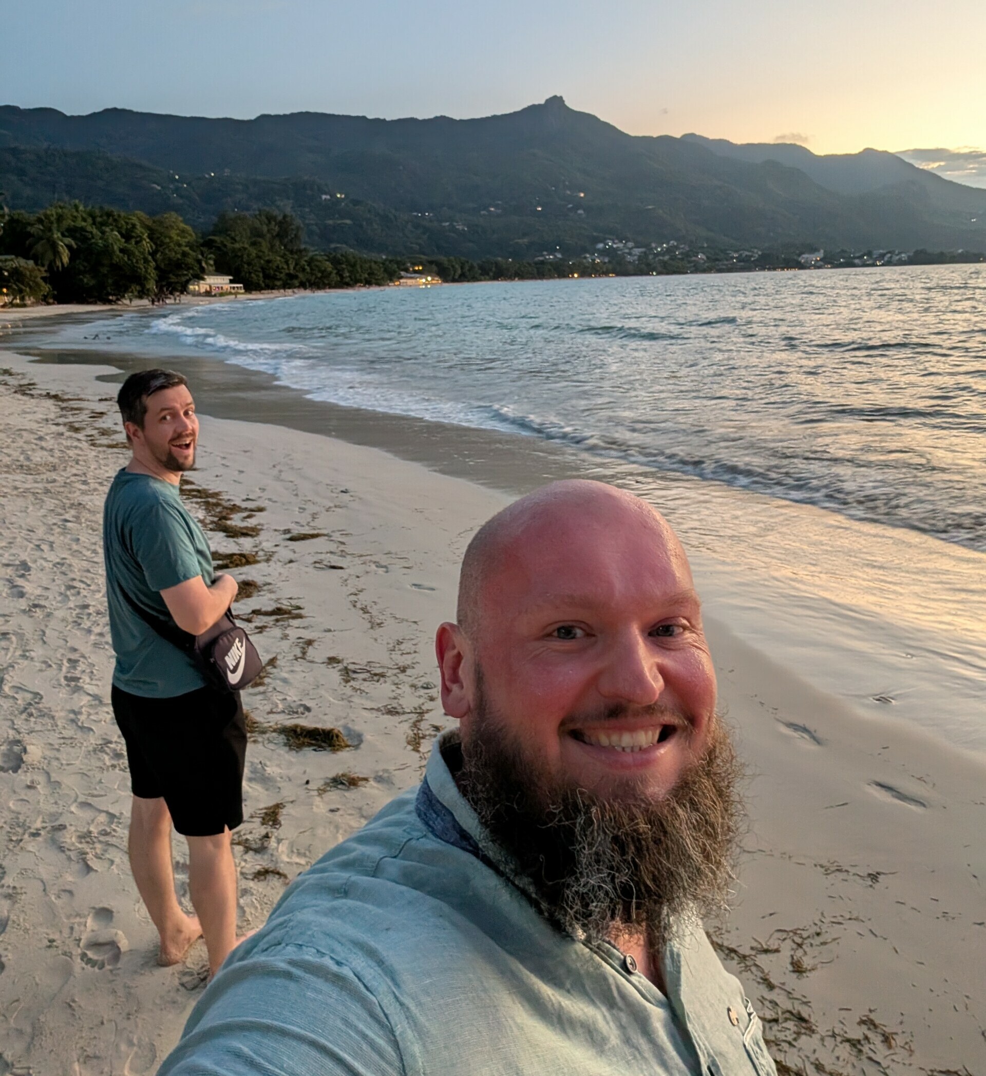 Siamar-Jungs am Strand Zwei Männer genießen einen sonnigen Strandspaziergang; ein Mann macht ein Selfie, während der andere im Hintergrund lächelt. Berge und der Ozean verschönern die Szenerie im Hintergrund.