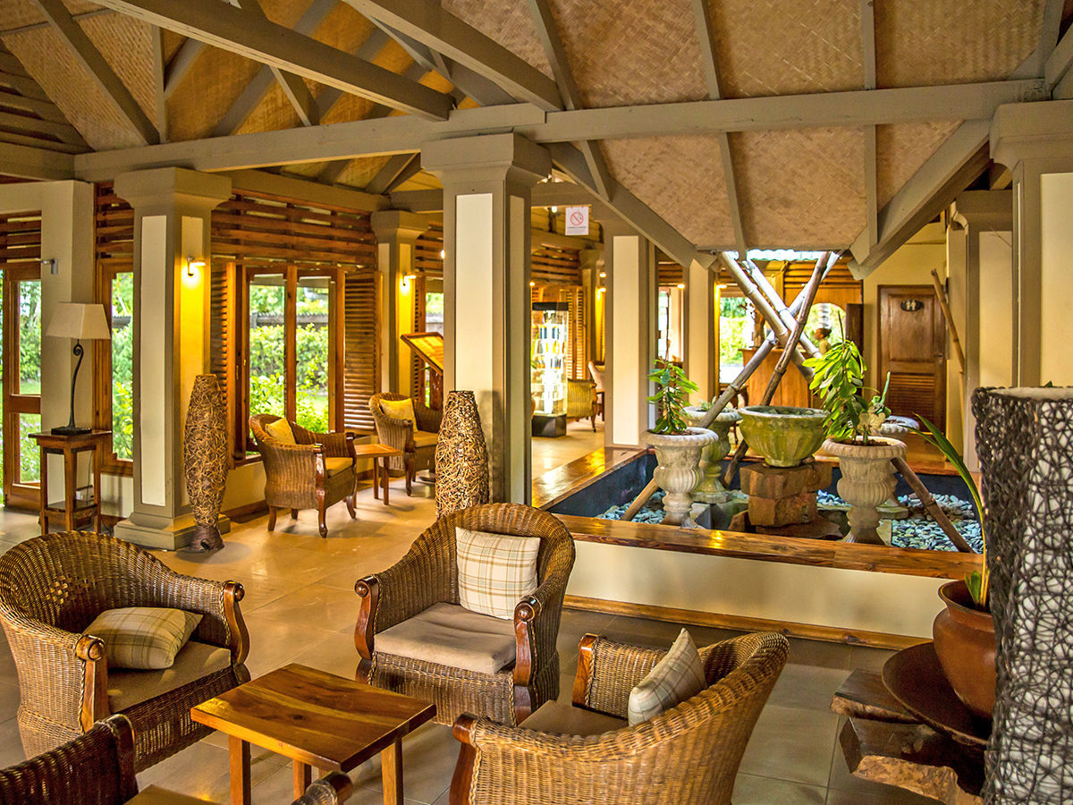Restaurant der Indian Ocean Lodge Ein elegantes und einladendes Innenraumdesign eines tropischen Hauses mit einer Kombination aus rustikaler und moderner Ästhetik, Holzmöbeln im Rattanstil und dekorativen Pflanzenarrangements. Der Raum ist mit Holzsäulen und Balken sowie zahlreichen Fenstern ausgestattet, die für eine helle und luftige Atmosphäre sorgen. Ein kleiner Indoor-Wasserbereich mit Steinelementen fügt ein ruhiges, natürliches Element hinzu.
