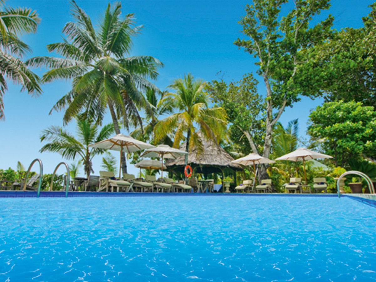 Pool der Indian Ocean Lodge Einladendes tropisches Resort mit einem großen, blauen Pool im Vordergrund, umgeben von hohen Palmen und grüner Vegetation. Am Rand des Pools stehen komfortable Liegestühle unter Sonnenschirmen.
