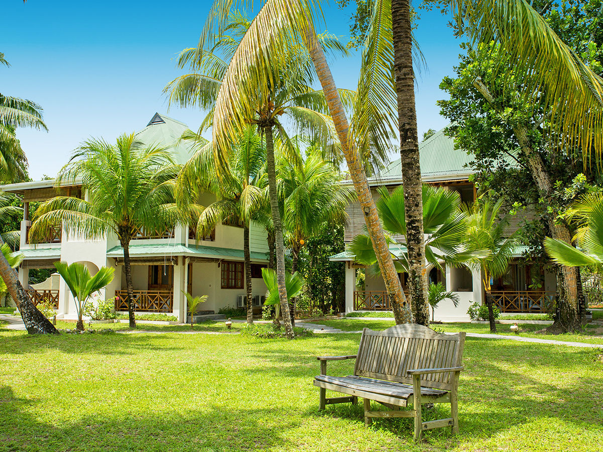 Garten der Indian Ocean Lodge Ein idyllisches tropisches Resort umgeben von üppigem Grün und hohen Palmen. Im Vordergrund befindet sich eine Holzbank auf einem gepflegten Rasen, während im Hintergrund stilvolle Bungalows mit weißen Säulen und Balkonen zu sehen sind. Helles Sonnenlicht taucht die Szenerie in warmes Licht.