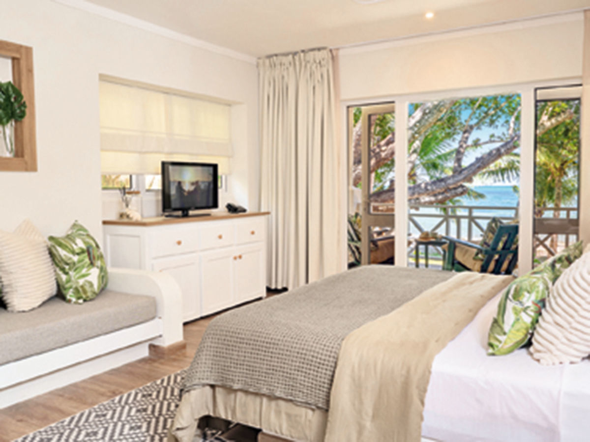 Zimmer mit Meerblick in der Indian Ocean Lodge Ein helles, gemütlich eingerichtetes Schlafzimmer mit einem Doppelbett, einer gepolsterten Bank und einer weißen Kommode mit Fernseher. Große Fenster bieten einen beeindruckenden Blick auf einen tropischen Strand mit Palmen.