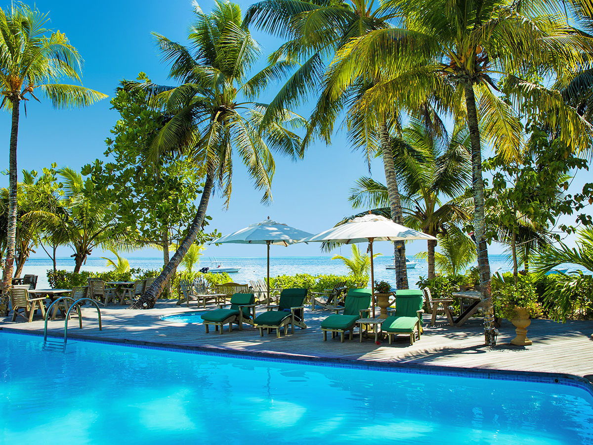 Pool mit Meerblick Alt-Text: Einladender Poolbereich in einem tropischen Resort, umgeben von üppigen Palmen und einer Vielzahl von Grünpflanzen. Im Vordergrund sieht man einen klaren blauen Pool mit Liegestühlen und Sonnenschirmen am Rand. Im Hintergrund erstreckt sich ein ruhiger Strand mit kristallklarem Meer und einem kleinen Boot, das nahe dem Ufer schwimmt.