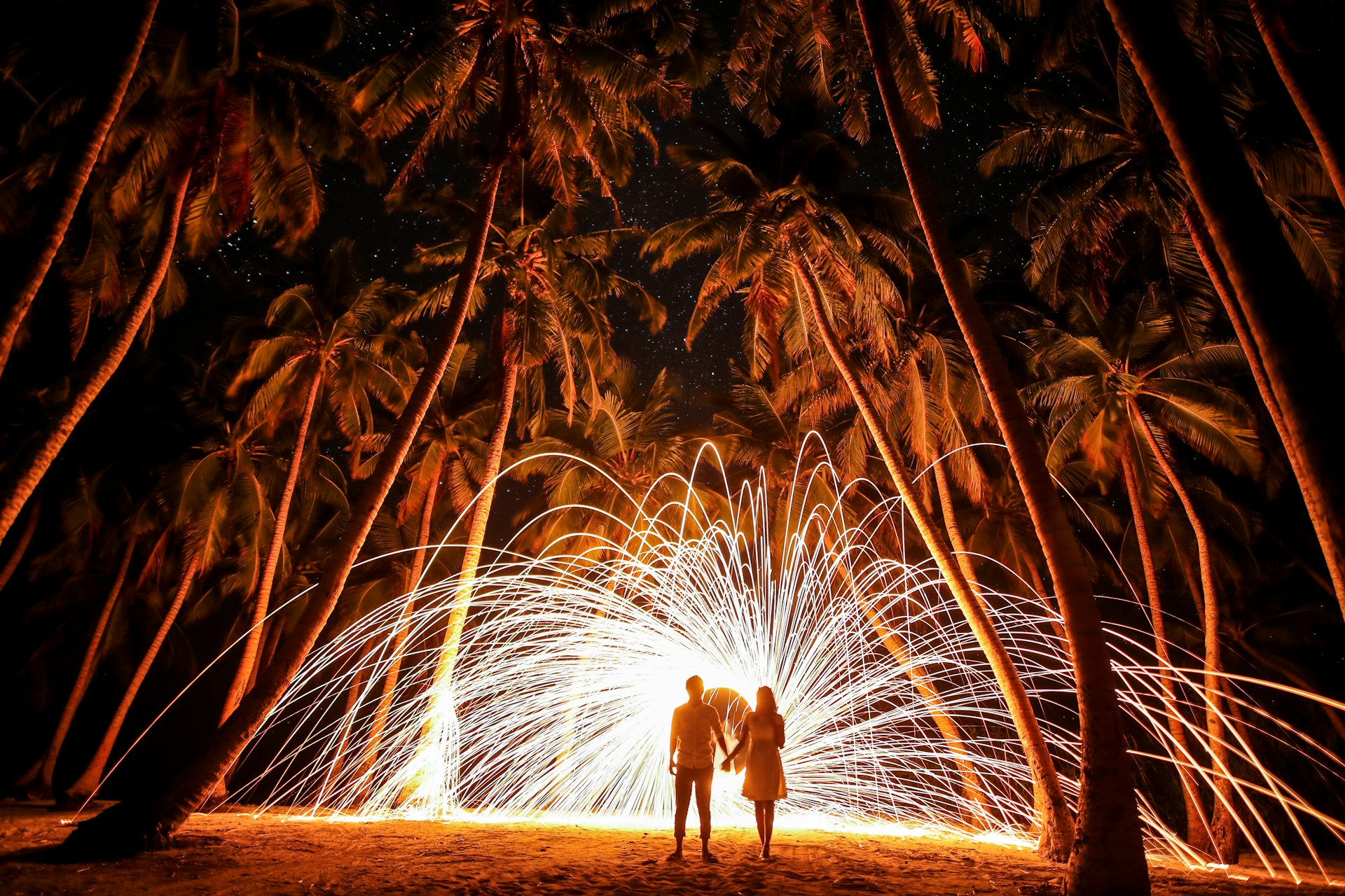 Feuerwerk unter Palmen Bild von einem Paar, das an einem nächtlichen Strand unter Palmen steht und einer spektakulären Feuerwerk-Show zusieht, wobei Funken in einem weiten Bogen durch die Luft fliegen, was eine spektakuläre Szene unter einem sternklaren Himmel kreiert.
