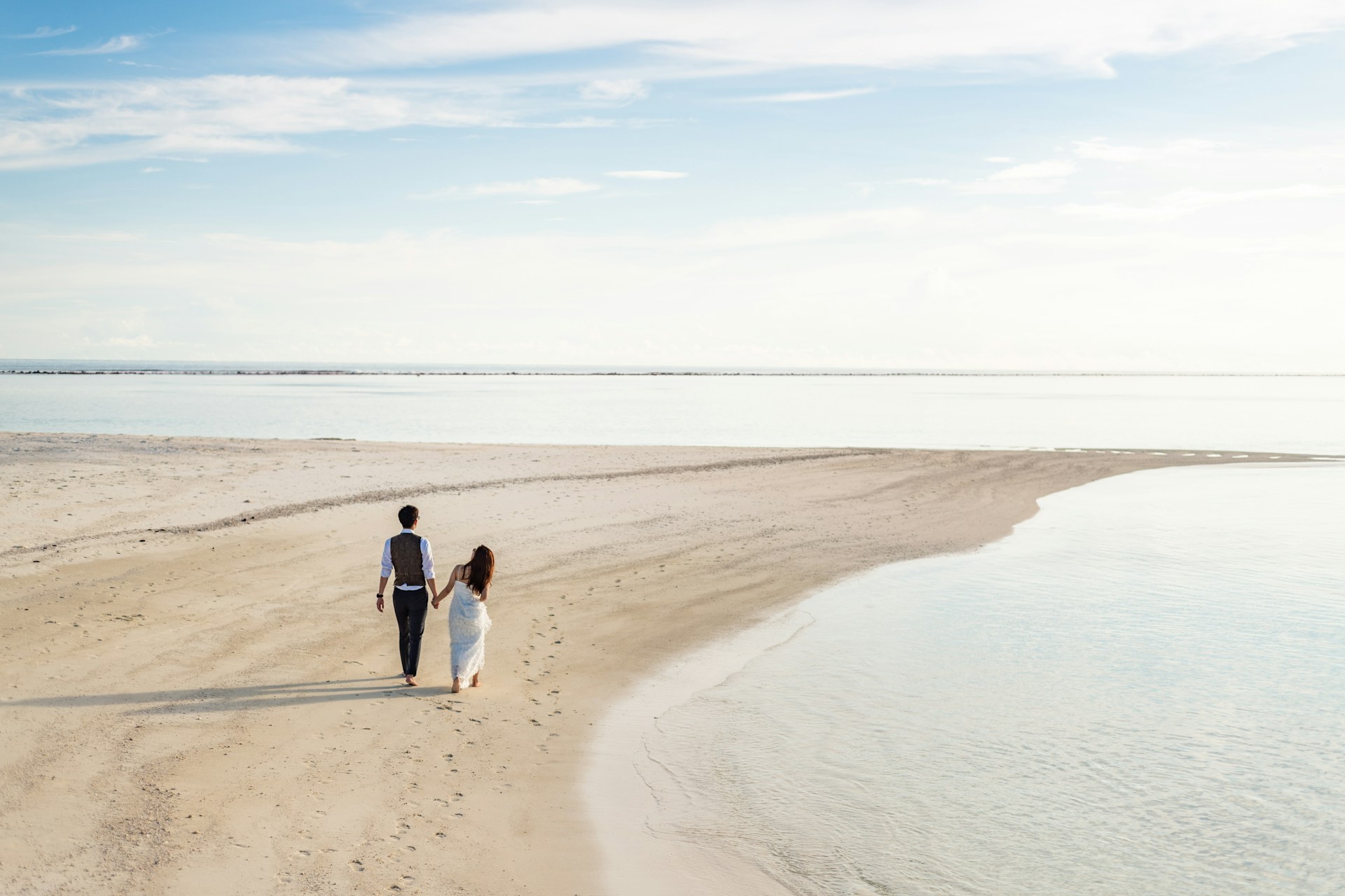Ruhige Meeresküste Ein frisch verheiratetes Paar spaziert Hand in Hand am Strand entlang. Die Szene zeigt eine ruhige Meeresküste mit feinem Sand und flachem Wasser. Der Himmel ist klar und blau mit einigen zarten Wolken, und die Atmosphäre vermittelt eine friedliche Stimmung.