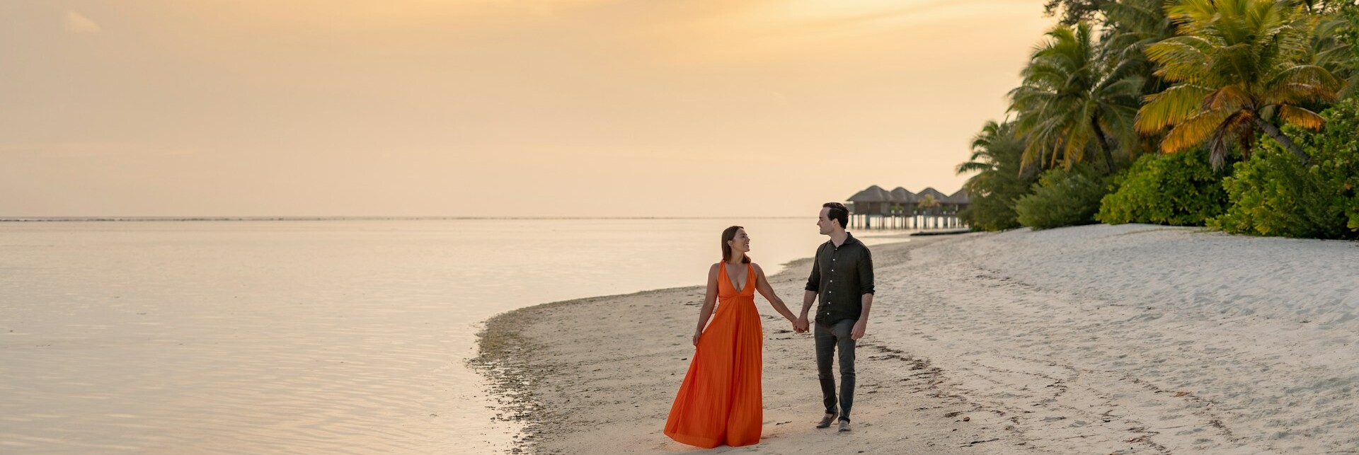 Spaziergang am Strand Ein Paar spaziert Hand in Hand am Strand entlang bei Sonnenuntergang. Die Frau trägt ein langes oranges Kleid und der Mann ist in einem leichten, dunklen Hemd und dunklen Hosen gekleidet. Der Strand ist von sanftem, weißem Sand und üppigem, grünem Laub umgeben, während im Hintergrund kleine Überwasser-Bungalows zu sehen sind. Der Himmel ist in warme Orange- und Gelbtöne getaucht, die das ruhige, spiegelglatte Meer reflektieren.
